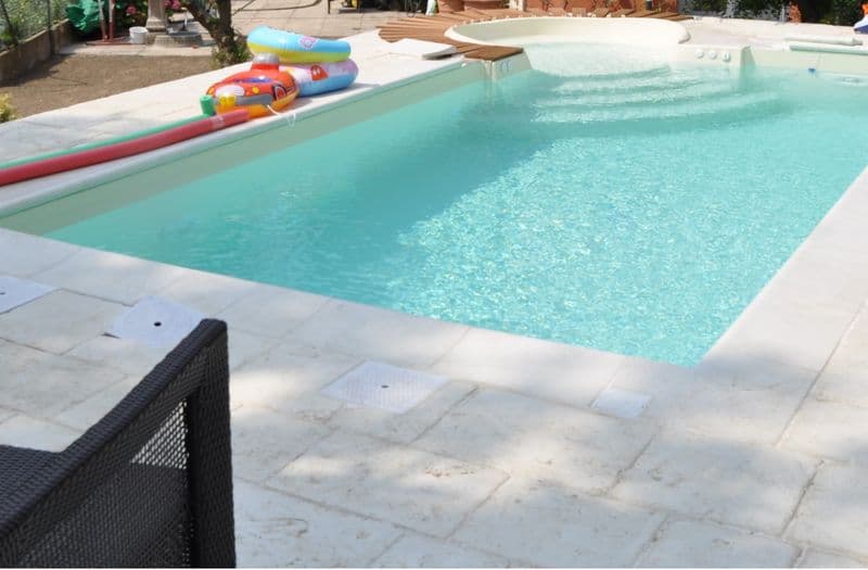 Piscina prefabbricata ultimata con bordo vasca in pietra chiara, seduta idromassaggio integrata e giardino