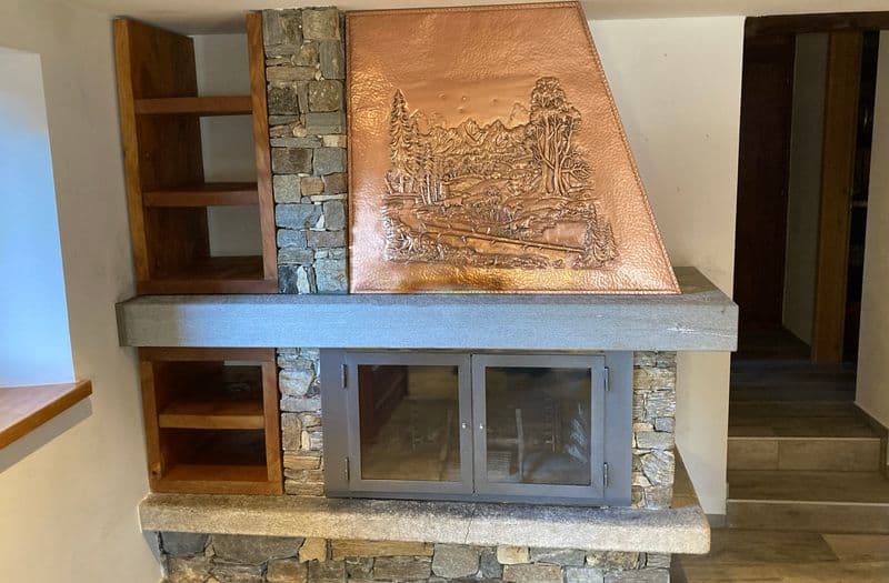Caminetto con cappa in rame sbalzato a motivo di paesaggio alpino, rivestimento in pietra e mensole in legno